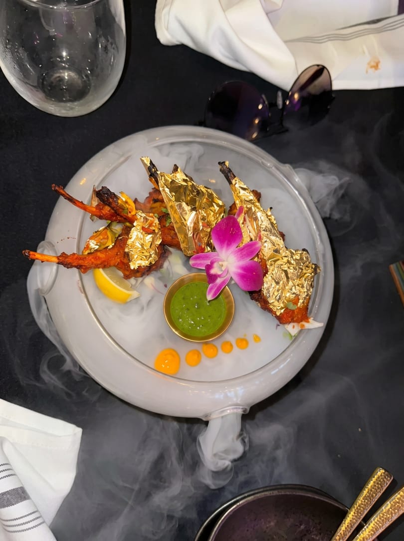 Gold-leafed chicken lollipops with green chutney and an orchid, served on a smoky platter.