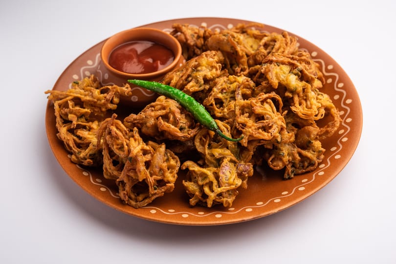 Crispy golden onion bhaji pakora with a side of red sauce and a green chili on a brown plate.