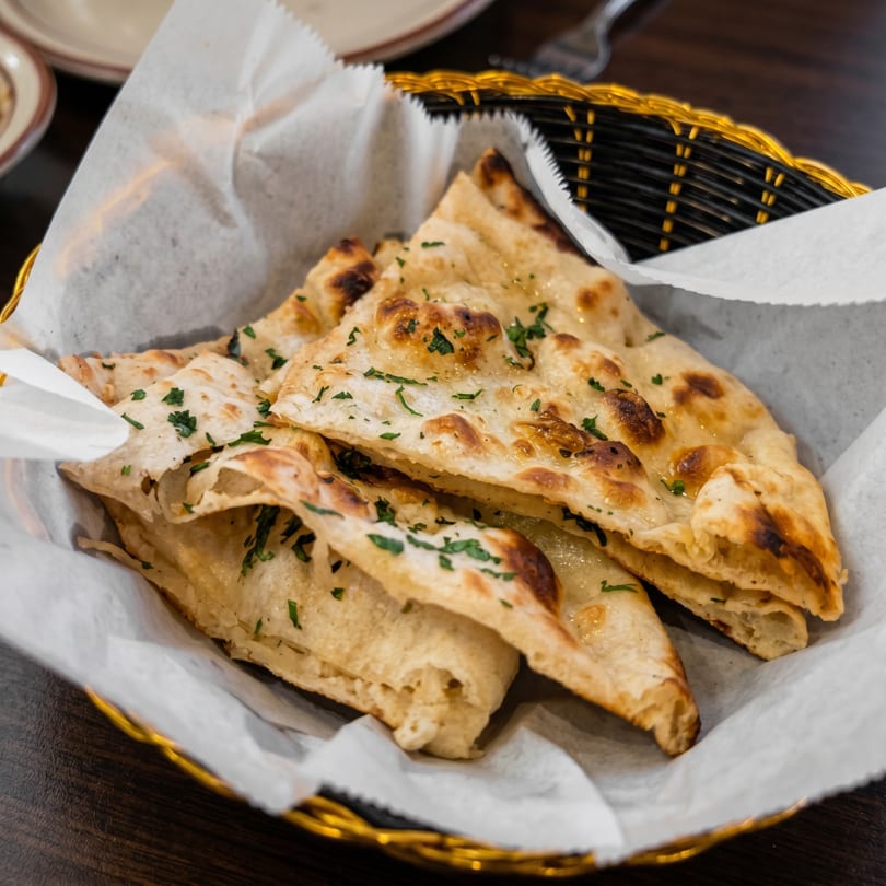 Golden-brown naan bread, garnished with fresh parsley, served in a basket lined with white paper.